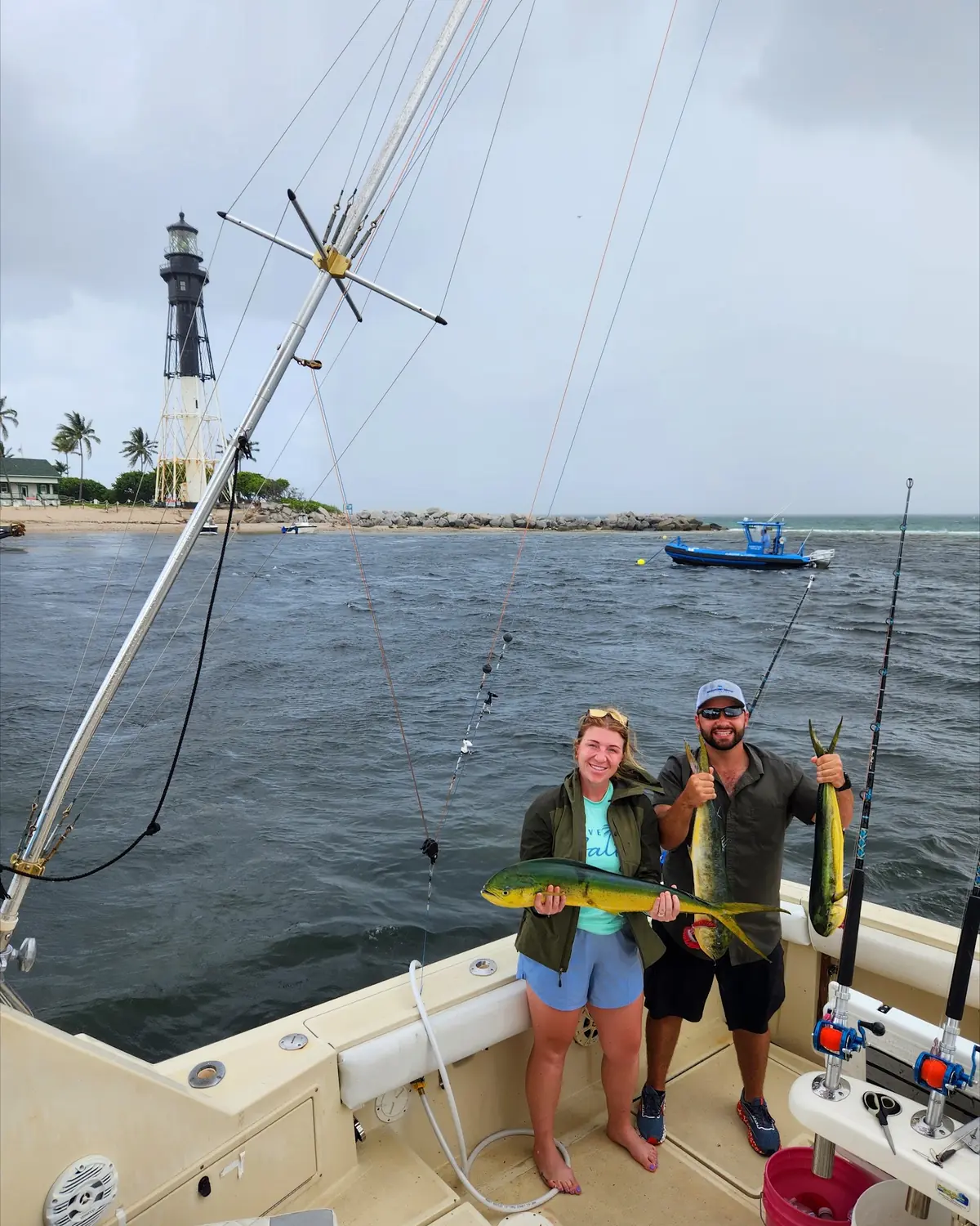 A couple holding up mahi mahi with the Hillsboro Inlet lighthouse behind the boat